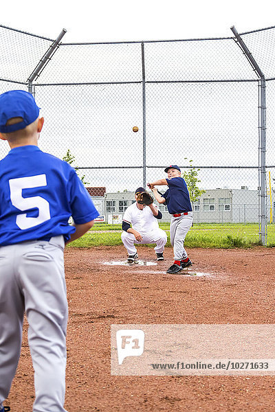 Ein Junge in einem Baseballtrikot wirft dem Schlagmann seiner Mannschaft den Ball zu  während sein Vater im Baseballdiamanten den Fänger spielt; Fort McMurray  Alberta  Kanada'.