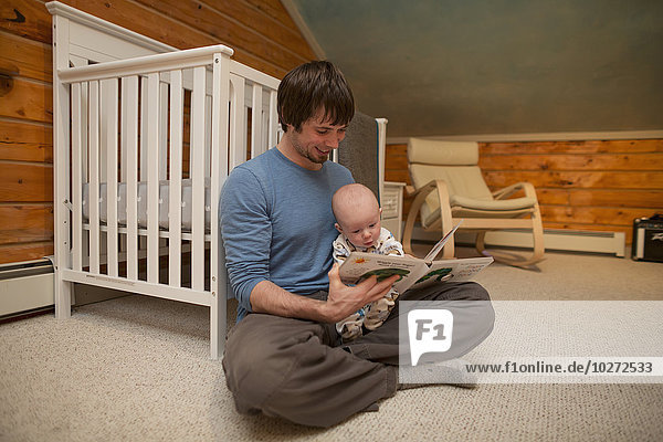 Vater hält sein Kind und liest ihm eine Gutenachtgeschichte vor  während er im Kinderzimmer auf dem Boden sitzt  USA