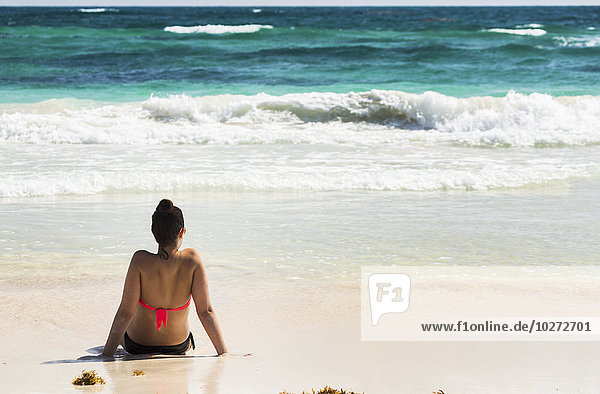 'Young woman sitting on sandy beach in water looking out at waves coming in and blue sky; Akumal  Quintana Roo  Mexico'