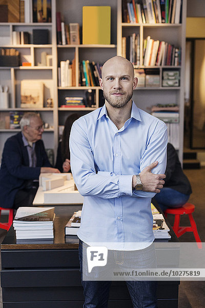 Portrait of confident businessman standing arms crossed while colleagues working in background at office