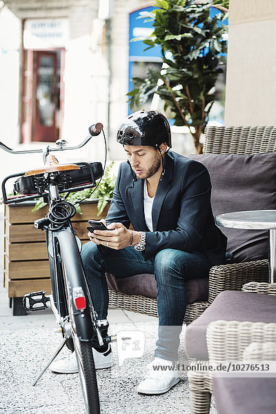 Geschäftsmann mit Fahrrad mit Smartphone beim Sitzen auf der Couch im Straßencafé