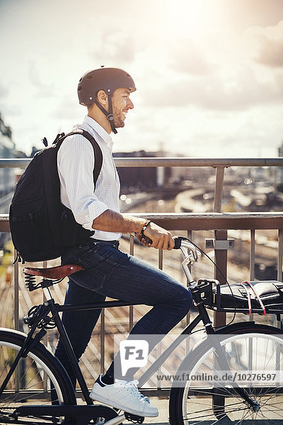 Lächelnder Geschäftsmann mit Fahrrad und Blick auf die Stadt  während er auf der Brücke steht.