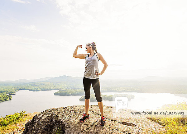 Junge Frau steht auf dem Gipfel eines Berges und spannt Muskeln an