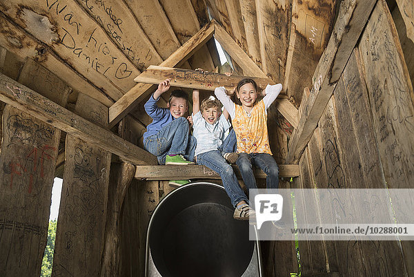 Drei Kinder sitzen auf dem Holzbalken eines Baumhauses auf einem Spielplatz  München  Bayern  Deutschland