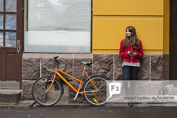 Finnland Helsinki Berghall Junge Frau mit orangefarbenem Fahrrad an der Gebäudewand lehnend