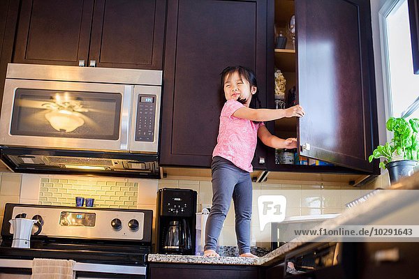 Junges Mädchen steht auf Küchenarbeitsplatte und sucht Leckerbissen im Schrank