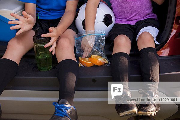 Taille unten des Jungen und der jüngeren Schwester  die in der Fußballtrainingspause im Kofferraum sitzen und Orangen essen.