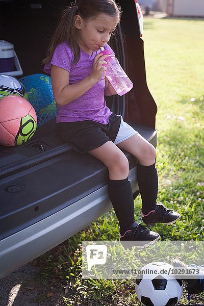 Mädchen-Fußballspielerin sitzt im Kofferraum und trinkt Wasser.