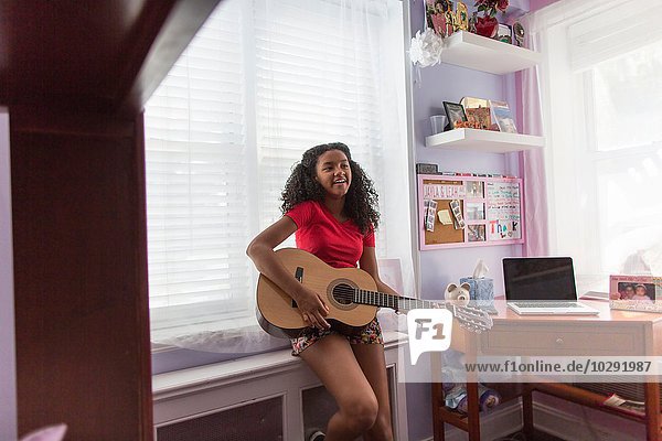 Mädchen lehnt sich gegen die Fensterbank  spielt Gitarre und schaut lächelnd weg.