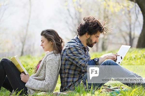 Young couple back to back on picnic blanket using digital tablet and reading