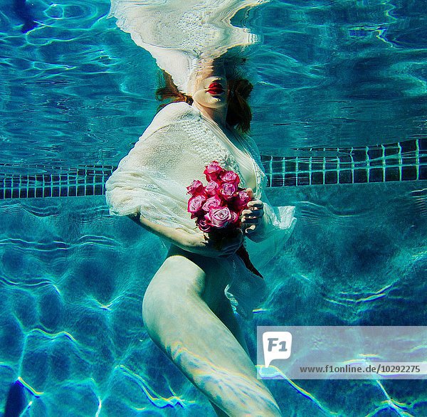 Junge Frau unter Wasser  trägt dünnes weißes Hemd und hält Blumen.