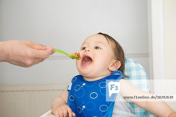 Hand of mother feeding baby food to open mouthed son in kitchen high chair