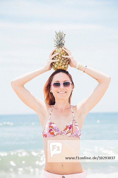 Porträt einer mittleren erwachsenen Frau am Strand  mit Sonnenbrille und Ananas am Kopf