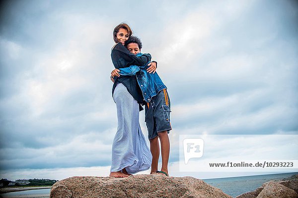 Portrait of mature woman and son hugging on coastal rocks  Cape Cod  USA