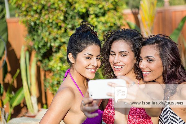 Three adult sisters wearing bikini tops posing for smartphone selfie in garden