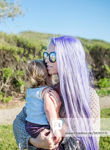 Junge Frau mit langen violetten Haaren  die eine kleine Tochter hält.