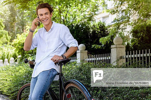 Junger Mann  der sich mit dem Smartphone gegen das Fahrrad lehnt und lächelnd wegschaut.
