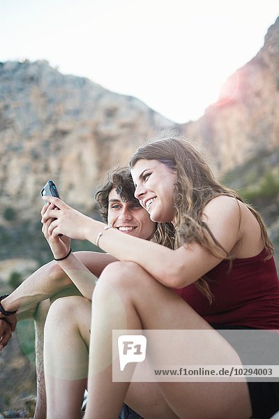 Junger Mann und jugendliche Schwester beim Lesen von Smartphone-Texten am felsigen Strand  Javea  Spanien