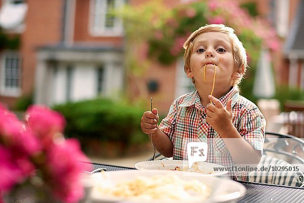 Junge sitzt am Gartentisch und saugt Spaghetti in die Kamera.
