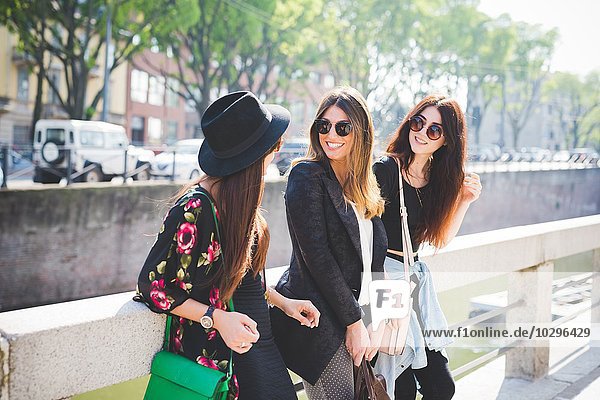 Three stylish young female friends chatting on city riverside
