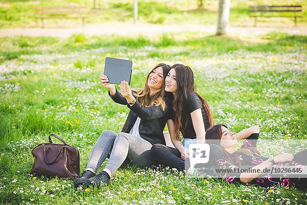 Young female friends sitting in park taking digital tablet selfie