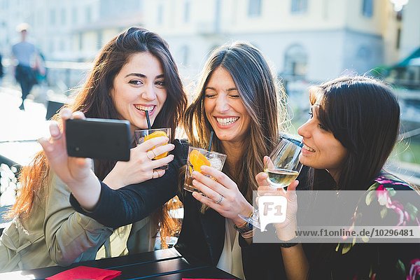 Three young women giggling for smartphone selfie at waterfront cafe