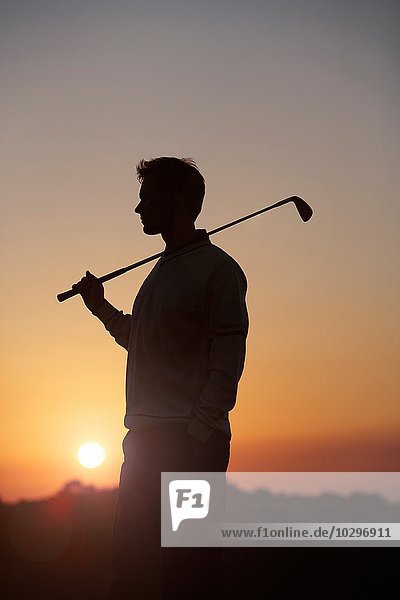 Golfer mit Golfschläger über der Schulter vor dem Sonnenuntergang  mit Blick nach vorne.