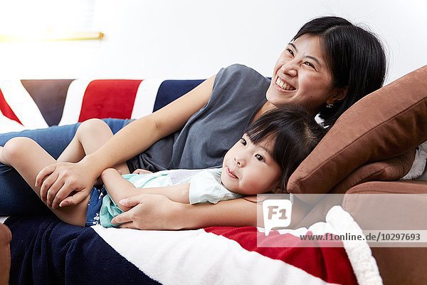 Junge chinesische Mutter und Tochter liegen auf dem Sofa und schauen gemeinsam zu Hause fernsehen.