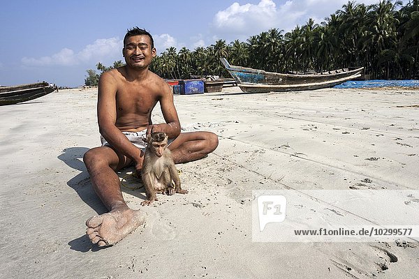 Einheimischer Mann sitzt mit einem Affen an der Leine am Strand des Fischerdorfes Ngapali  hinten Fischerboote  Thandwe  Rakhine-Staat  Myanmar  Asien