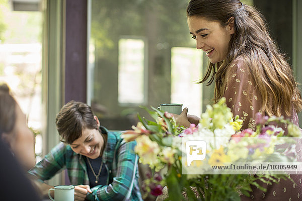 Drei sprechende und lachende Frauen  die um einen Tisch saßen. Großer Blumenstrauß in einer Vase.