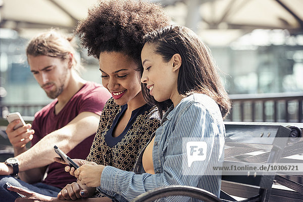 Three people seated side by side on a park bench checking their smart phones