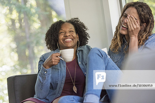 Zwei Frauen sitzen gemeinsam lachend auf einem Sofa  die eine hält eine große Kaffeetasse.