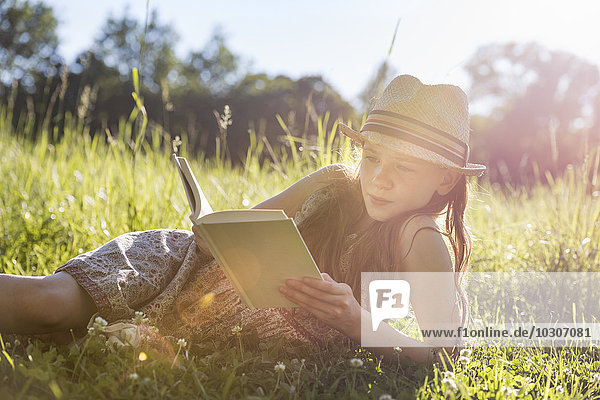 Ein junges Mädchen mit einem Strohhut  das im Gras liegt und ein Buch liest.