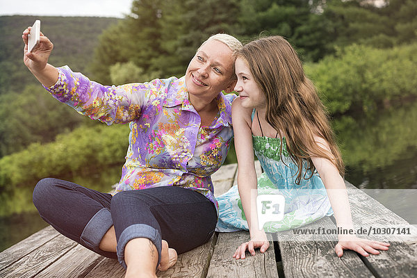 Eine Frau und ein Kind sitzen auf einer Anlegestelle am See und machen einen Selfie mit einem Smartphone.