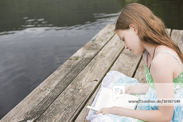 Ein junges Mädchen sitzt auf dem Steg an einem See und liest.