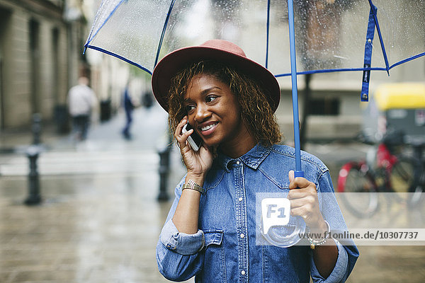 Spanien  Barcelona  Portrait einer lächelnden jungen Frau mit Schirm und Smartphone