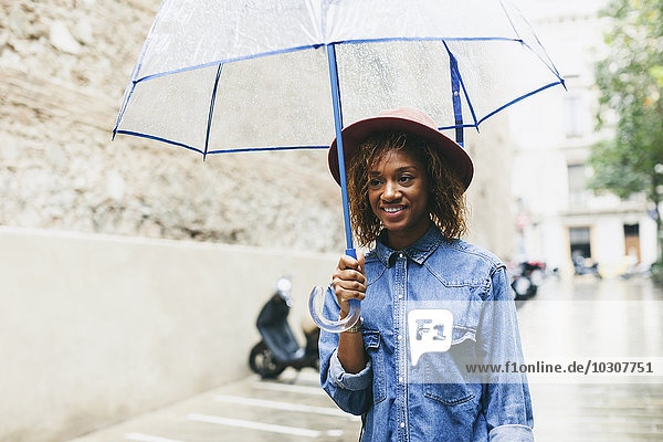 Spanien  Barcelona  Porträt einer lächelnden jungen Frau mit Regenschirm in Hut und Jeanshemd