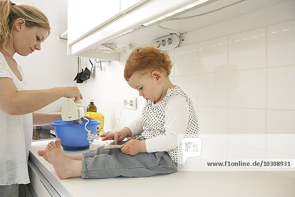 Kleines Mädchen sitzt auf der Küchentheke mit Smartphone  während ihre Mutter backt