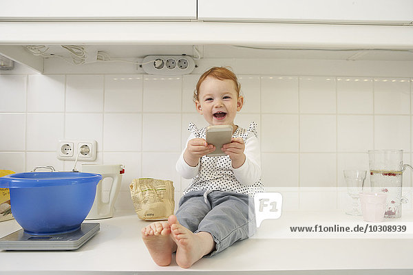 Lachendes kleines Mädchen sitzend auf der Küchenzeile mit Smartphone