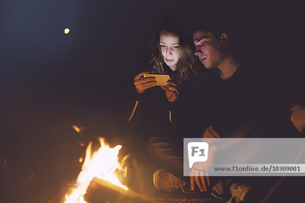 Young couple sitting together at the campfire looking at cell phone