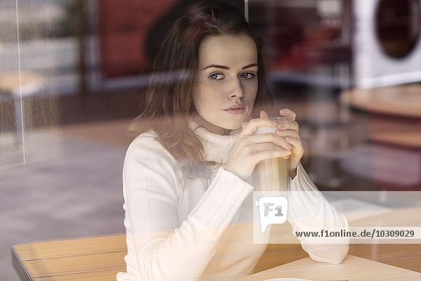 Porträt einer jungen Frau mit Latte Macchiato in einem Café mit Blick durch die Fensterscheibe