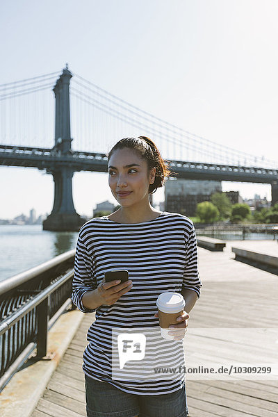USA  New York City  portrait of smiling young woman with smartphone and coffee to go