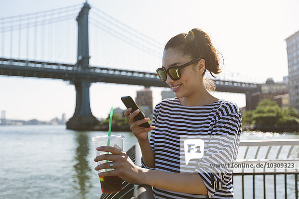 USA  New York City  portrait of smiling young woman with soft drink looking at smartphone