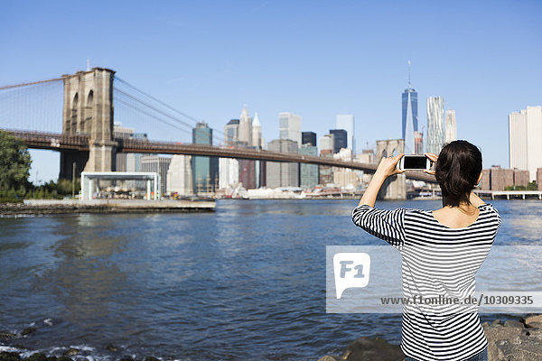 USA  New York City  back view of young woman taking a photo of Brooklyn Bridge with smartphone