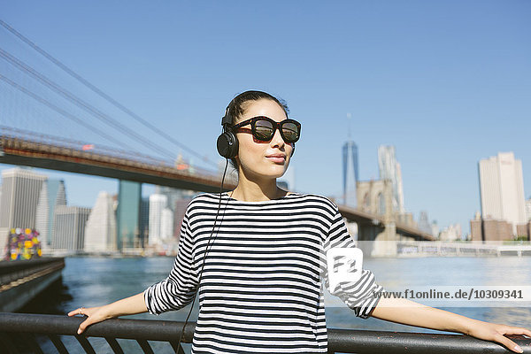 USA  New York City  Portrait einer jungen Frau mit Kopfhörer und Sonnenbrille vor der Skyline