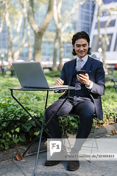 USA  New York City  Manhattan  smiling businessman with laptop and cell phone in Bryant Park