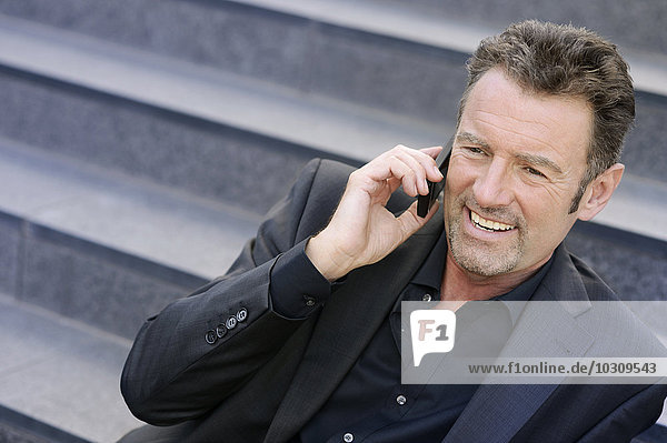 Porträt eines lächelnden Geschäftsmannes  der auf einer Treppe sitzt und mit dem Smartphone telefoniert.