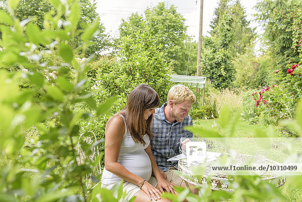 Young couple expecting baby  sitting in garden reading magazine