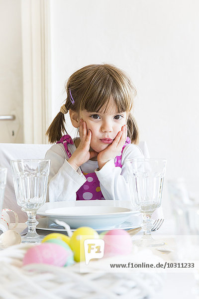 Porträt eines kleinen Mädchens mit Kopf in der Hand,  das am gedeckten Esstisch sitzt.