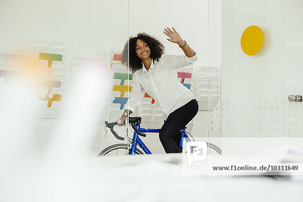 Lächelnde junge Frau mit Fahrrad im Büro winkend